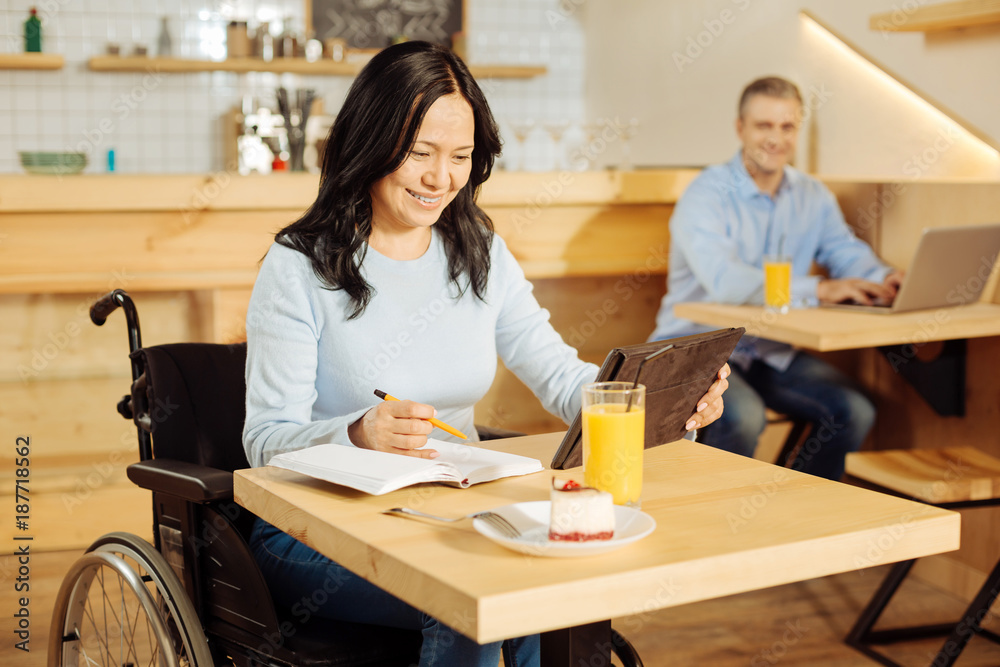 Good mood. Beautiful cheerful disabled woman sitting in a wheelchair and writing in her notebook and working on her tablet in a cafe and a man sitting in the background Good mood. Beautiful cheerful disabled woman sitting in a wheelchair and writing in her notebook and working on her tablet in a cafe and a man sitting in the background