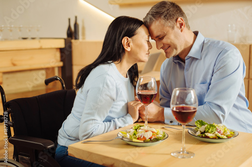 Romantic dinner. Beautiful delighted disabled woman and an attractive inspired well-built man sitting in a cafe with eyes closed and holding hands and having dinner
