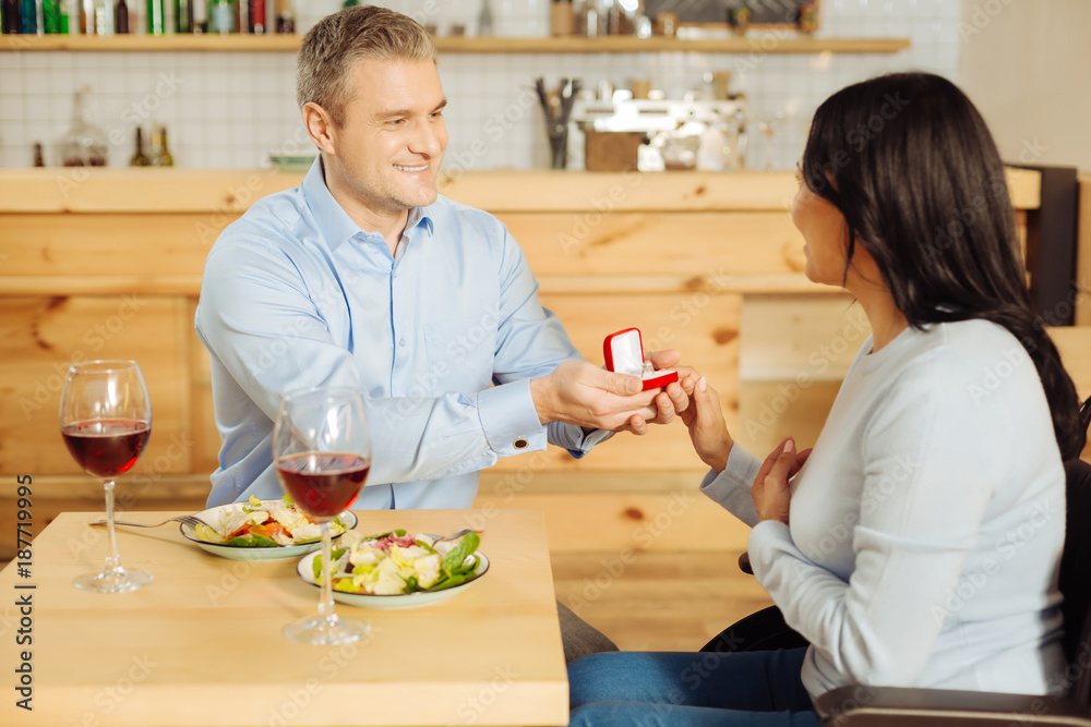 Bridegroom. Good-looking inspired well-built man smiling and proposing to his beautiful content handicapped woman while holding a ring and having dinner Bridegroom. Good-looking inspired well-built man smiling and proposing to his beautiful content handicapped woman while holding a ring and having dinner