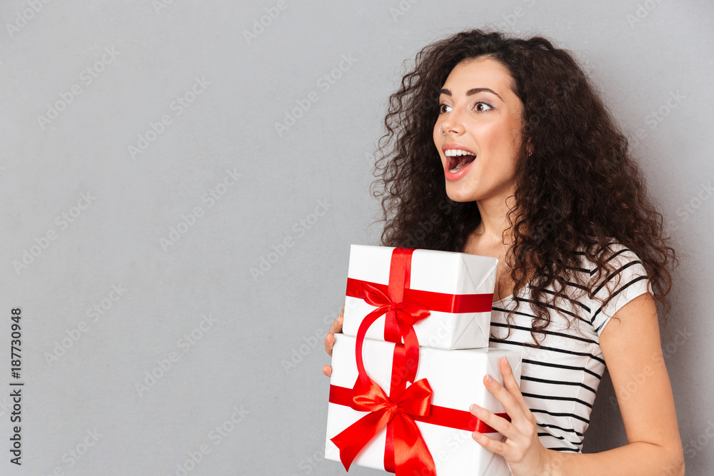 Happy emotions of beautiful woman in striped t-shirt holding two gift-wrapped boxes with red bows while standing over grey background