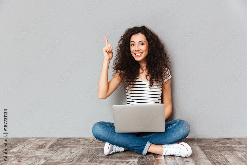 Fototapeta premium Nice woman with beautiful smile being excited to find useful information in internet via silver computer gesturing eureka sitting in lotus pose on the floor