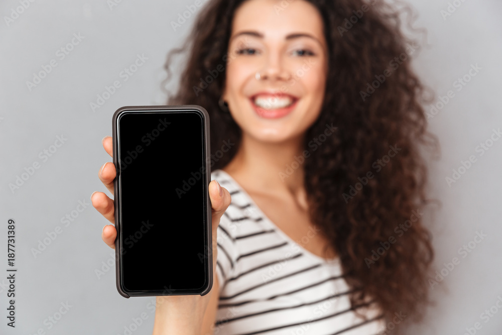 Attractive adult girl with ring in nose demonstrating her mobile phone on camera advertising new model being glad while isolated against grey wall Attractive adult girl with ring in nose demonstrating her mobile phone on camera advertising new model being glad while isolated against grey wall