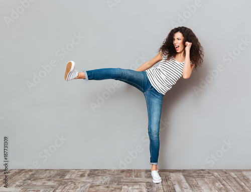 Portrait of strong young female with curly brown hair kicking invisible opponent punching with leg over grey wall photo