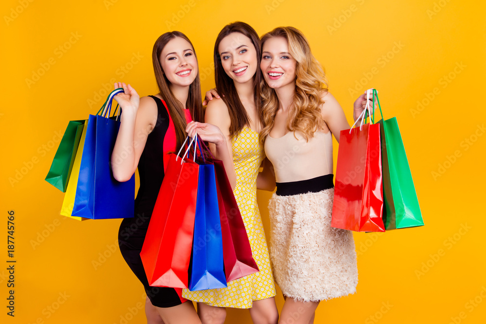 Three pretty, charming, successful girls in dresses holding colorful shopping bags, spent a lot of money, crazy sales, shopaholic, buyers, credit card, yellow background