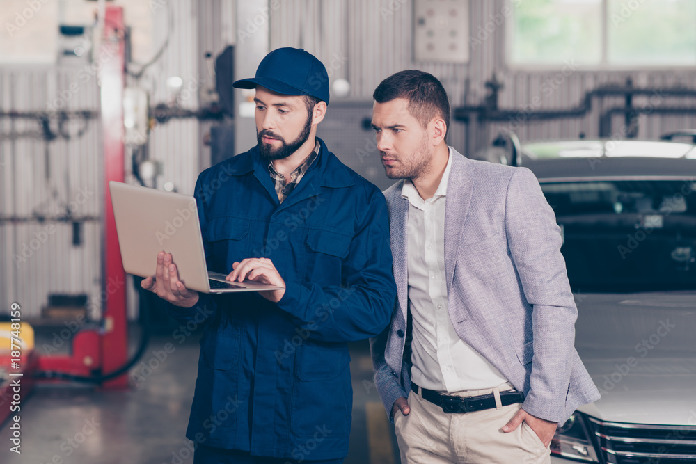 Experienced  bearded  specialist in cap demonstrating the damages of businessman`s car, showing the way of car repairing to client  on his device in workstation