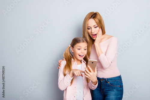 Attractive, shocked, impressed, comic kid pointing with index finger to smart phone screen to her amazed mother, showing something interesting, mom hold hand on cheek, together with open mouth