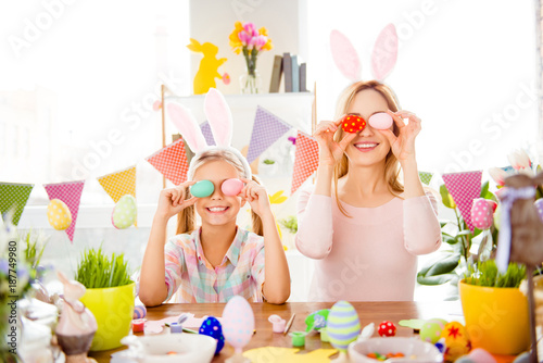 Little, positive, cheerful daughter and her mom, preparing for Easter, sitting at desk, holding colored, painted eggs on eyes place, enjoying seasonal spring holiday © deagreez