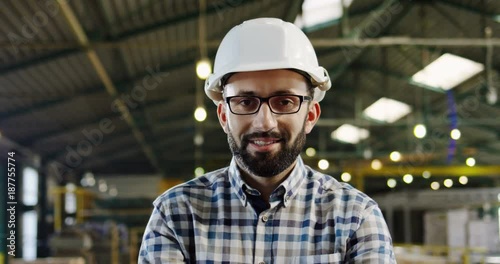 Close up of a man factory worker in a helmet, glasses and plaid shirt posing to the camera in a big empty factory werehouse. Inside. Portrait shot photo