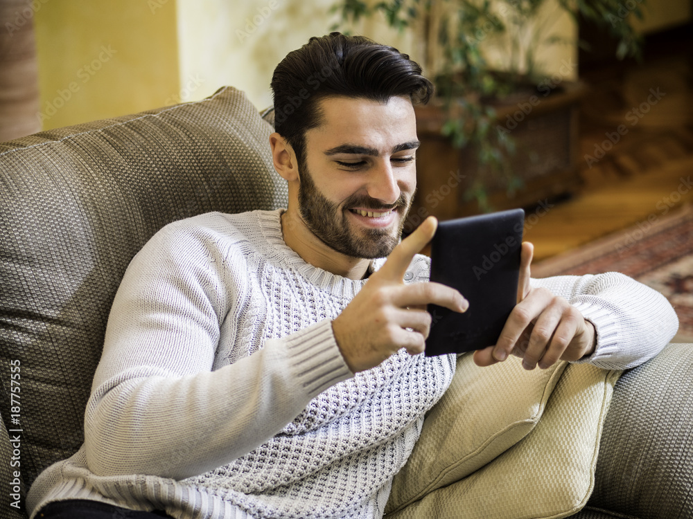 Handsome young man at home smiling, reading with ebook reader lying on a couch