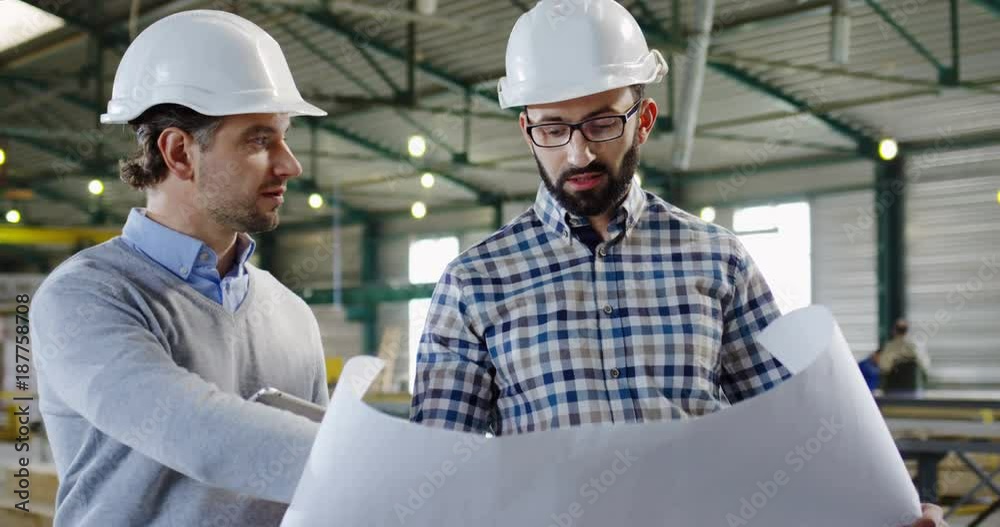 Two Caucasian handsome men in helmets talking and looking at the big paper with drawings and discussing it. Factory space. Indoor. Portrait. Close up Two Caucasian handsome men in helmets talking and looking at the big paper with drawings and discussing it. Factory space. Indoor. Portrait. Close up
