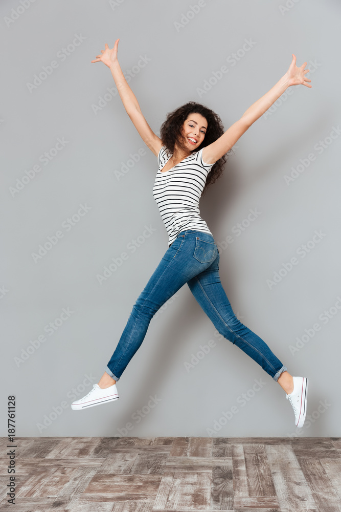 Energetic woman 20s in striped t-shirt and jeans jumping with hands throwing up in air over grey background