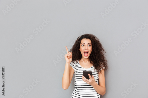Smart female with mobile phone in hand keeping index finger pointed upwards expressing she has an idea or saying eureka over grey background photo