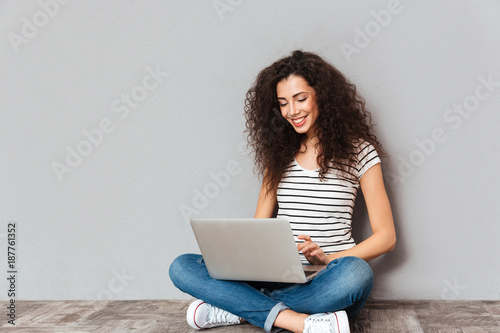 Attractive female with beautiful smile interacting in skype or typing message using silver notebook while sitting in lotus pose on the floor over grey wall