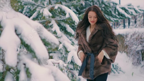 Close-up of Brunette fashionable sexy pretty rich woman in brown fur coat brushes woman walking in winter time slow motion. Happy girl having fun in snow. Snow covered Christmas trees in the winter