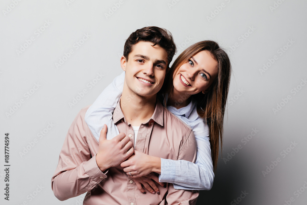 Portrait of a cheerful young couple hugging while standing and looking at camera over gray Portrait of a cheerful young couple hugging while standing and looking at camera over gray