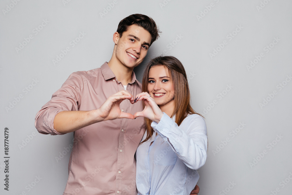 Two young smiling people in casual clothing hugging each other and making hear using their hands isolated on gray Two young smiling people in casual clothing hugging each other and making hear using their hands isolated on gray