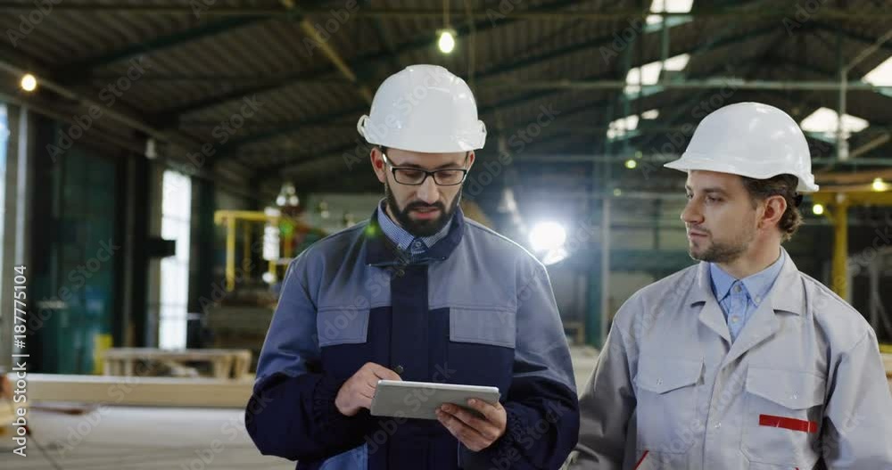 Two male engineers in helmets with a tablet device walking and discussing the new project inside a big furniture factory. Working process on the background. Portrait shot Two male engineers in helmets with a tablet device walking and discussing the new project inside a big furniture factory. Working process on the background. Portrait shot