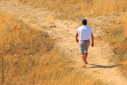 A man in shorts, a white T-shirt and a blue cap walking along a sandy and stony path among bushes, grass and flowers.