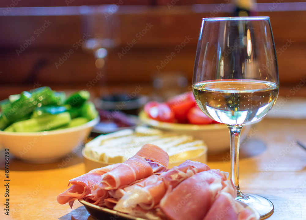 Sliced prosciutto with a glass of wine and vegetables in the background.