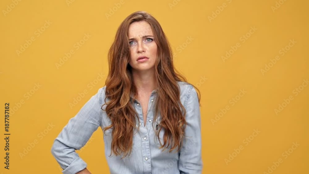 Displeased ginger woman in denim shirt gesturing with her finger against temple and looking at the camera over yellow background