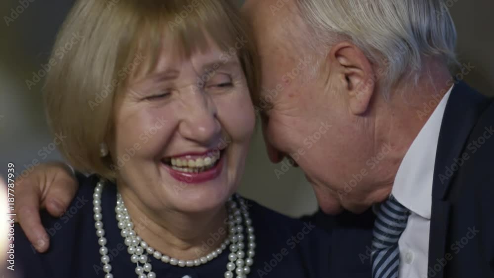 Medium shot of smiling elderly man in suit whispering something in ear of happy senior woman drinking wine on romantic date in restaurant Medium shot of smiling elderly man in suit whispering something in ear of happy senior woman drinking wine on romantic date in restaurant