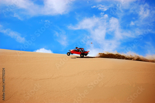 Foto Orange dune buggy tearing across golden sand dunes under a vivid blue sky, kicking up a dramatic spray of sand for an exhilarating off‑road desert adventure