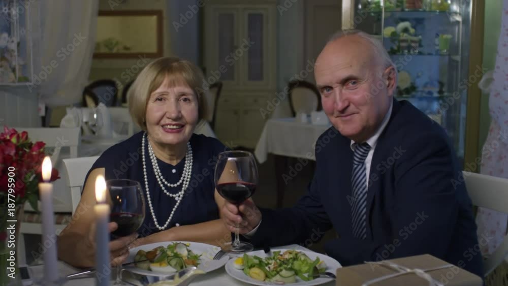 Zoom in of cheerful senior couple holding glasses with wine and looking at camera while having romantic date in restaurant Zoom in of cheerful senior couple holding glasses with wine and looking at camera while having romantic date in restaurant