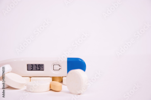 The electronic thermometer shows a high body temperature. White pills and orange antibiotics on a white background. Influenza and seasonal illnesses.