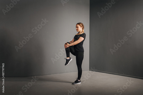Fitness runner woman doing warm-up routine in gym  stretching leg muscles with standing single knee to chest stretch. Female athlete preparing legs for cardio workout in sportswear.