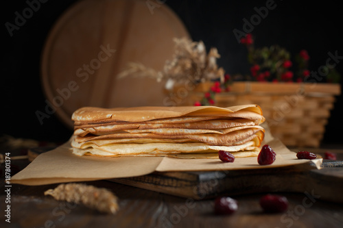 a pile of pancakes, spikelets, a round cutting board, a basket with branches with red berries and berries on a dark wooden table