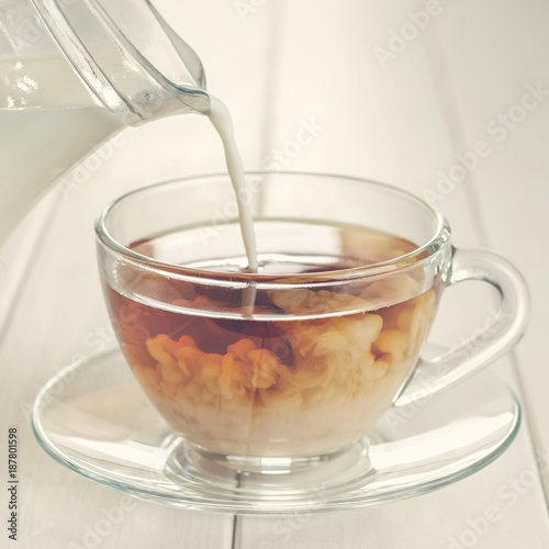 Pouring milk in tea in glass cup on white wooden table.