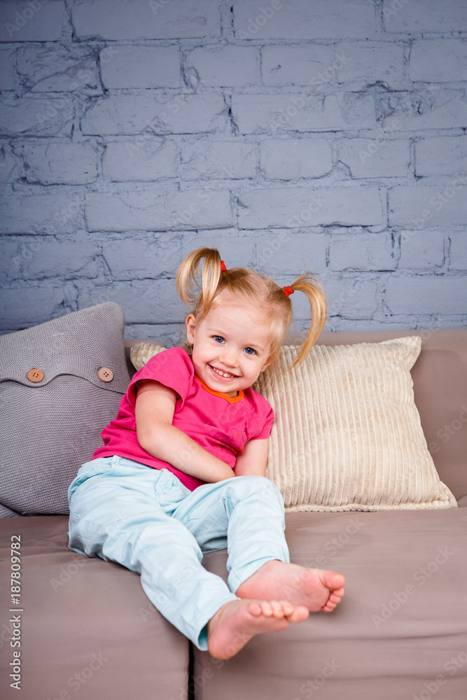 Little Funny Girl Pampering and Playing On The Sofa At Home Showing Naked Heels. He laughs showing his tongue and teeth. She is dressed in bright stylish clothes Little Funny Girl Pampering and Playing On The Sofa At Home Showing Naked Heels. He laughs showing his tongue and teeth. She is dressed in bright stylish clothes