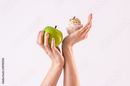 Try to choose. Studio shot of woman deciding between healthy and unhealthy food. Woman holding green apple in one hand and cake in other, isolated on white background. Healthy lifestyle concept