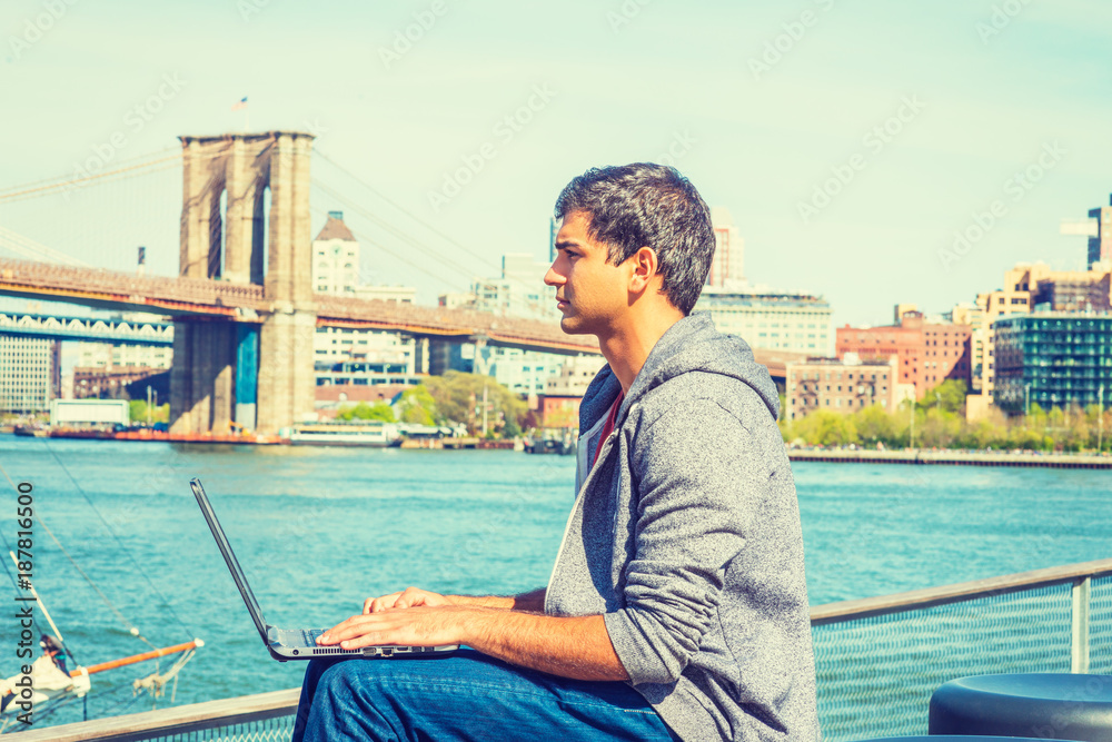 Way to Success.  East Indian American student travels, studies in New York, wearing hooded sweatshirt, jeans, sits by river, works on laptop computer, seriously thinks. Brooklyn bridge on background..
