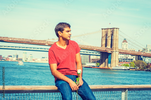 I miss you. Young East Indian American man wearing red V neck T shirt, jeans, sits on fence by river, holds white rose, looks around, thinks. Manhattan, Brooklyn bridges on background. Filtered effect