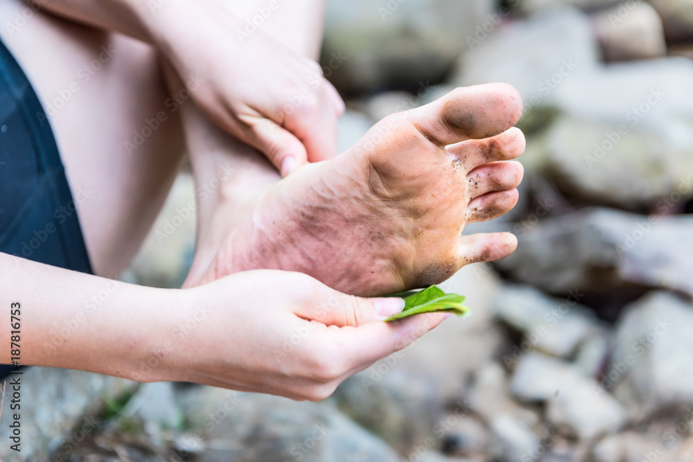 Closeup of young woman's dirty foot with her cleaning dirt with green leaf in nature by rocks