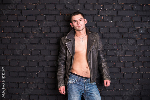 Attractive muscular sexual young man wearing denim trousers and a leather jacket. black brick wall on background © Serhii Barylo