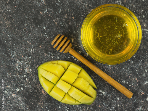 Cut mango and a bowl of honey and spoon on a dark stone table.