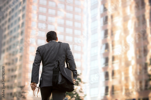Business man walking to work, motivational conceptual perspective from behind with buildings and copy space