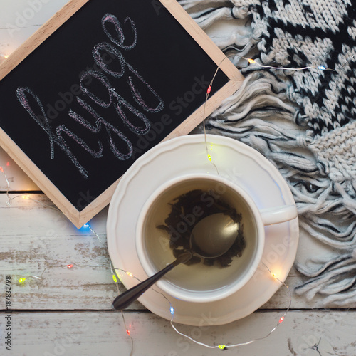 Square photo of green tea in small cup, knitted woolen blanket and blackboard with handwritten word "hygge" on wooden background, top view. Hygge concept. Comfort and coziness