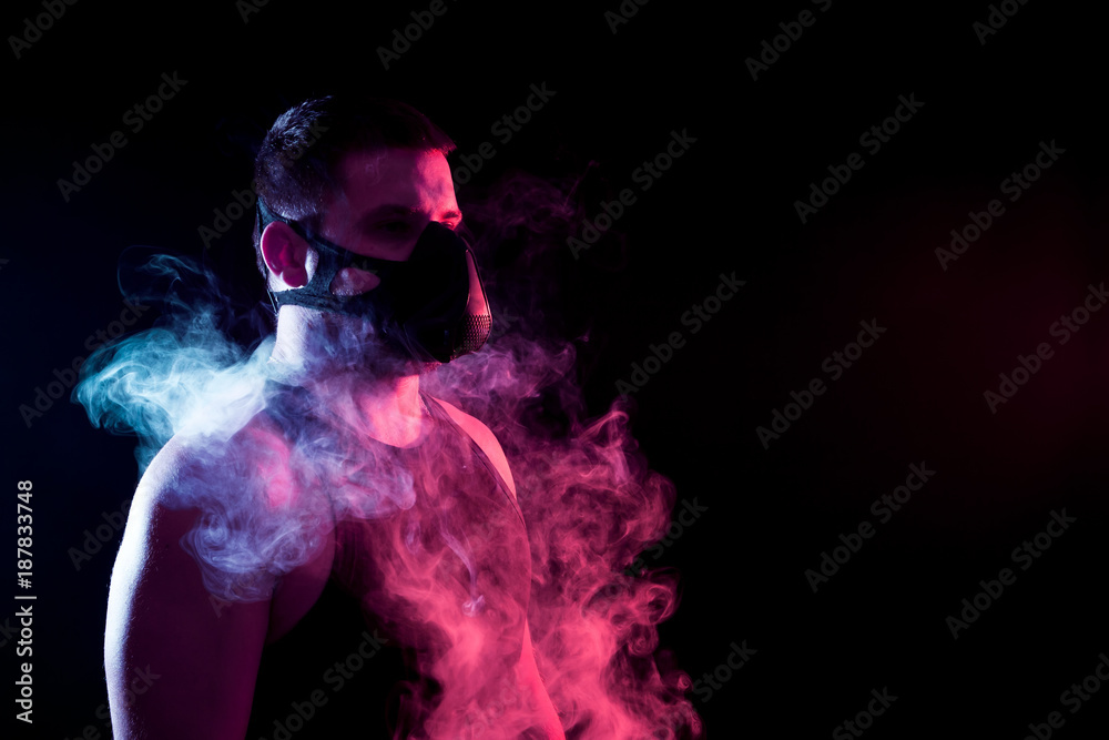 A dark-haired man breathes through a training black mask around a blue and red cloud of smoke from a wipe on a black isolated background