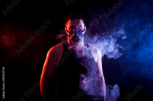 A young athletic man in a sports shirt breathes through a training black mask around a blue and red smoke cloud from a veip on a black isolated background