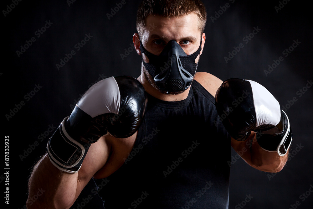 Portrait of young athletic man wearing  sport shirt, training mask and  boxing gloves on black isolated background
