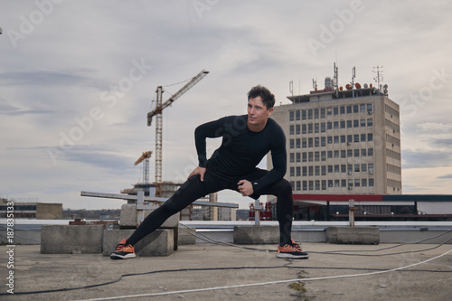 one young man stretching outdoors on rooftop. Buildings crane behind, cityscape, urban area.