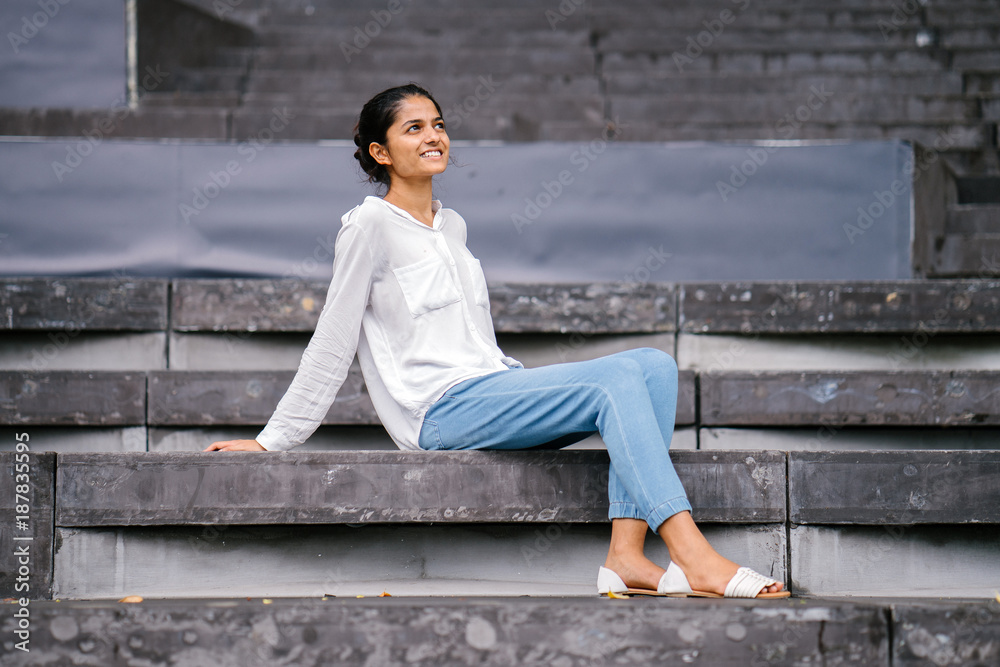 Portrait of attractive and young Indian woman sitting on grey steps and  smiling. She is well