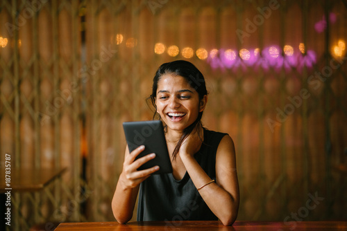 Portrait of young indian woman sitting at a cafe enjoying reading a kindle. The background is brown with lights. 