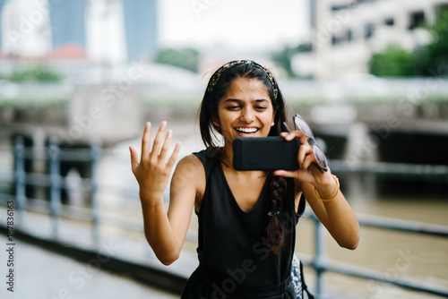 Young Indian woman smiling and beckoning to the viewer while she takes a photo on her smartphone. The Singapore River is in the background and it is in the daytime. 