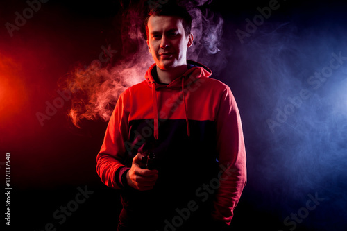 Man in sport hoody vaping an electronic cigarette.Isolated on black background.Around blue and red clouds of smoke