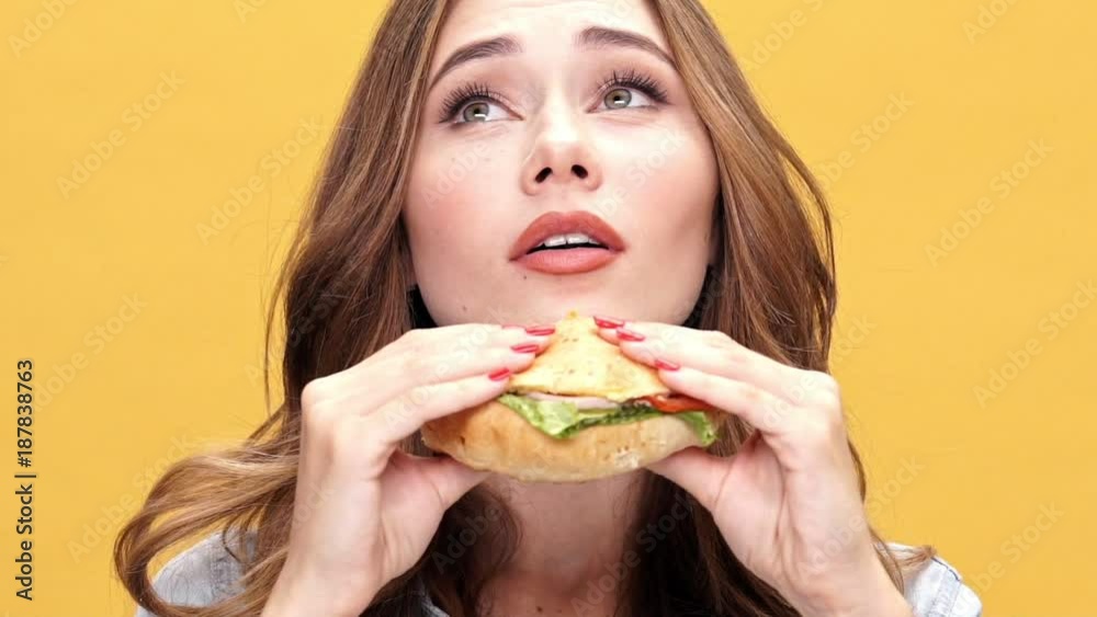 Close up view of Pleased brunette woman in denim shirt sniffing and enjoys of a big burger over yellow background