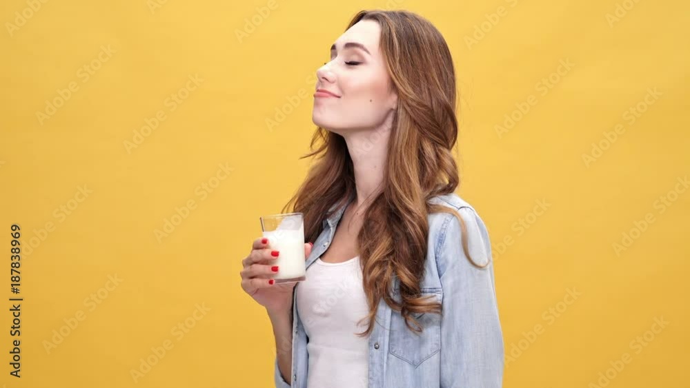 Happy brunette woman in denim shirt drinking kefir and looking at the camera over yellow background Happy brunette woman in denim shirt drinking kefir and looking at the camera over yellow background